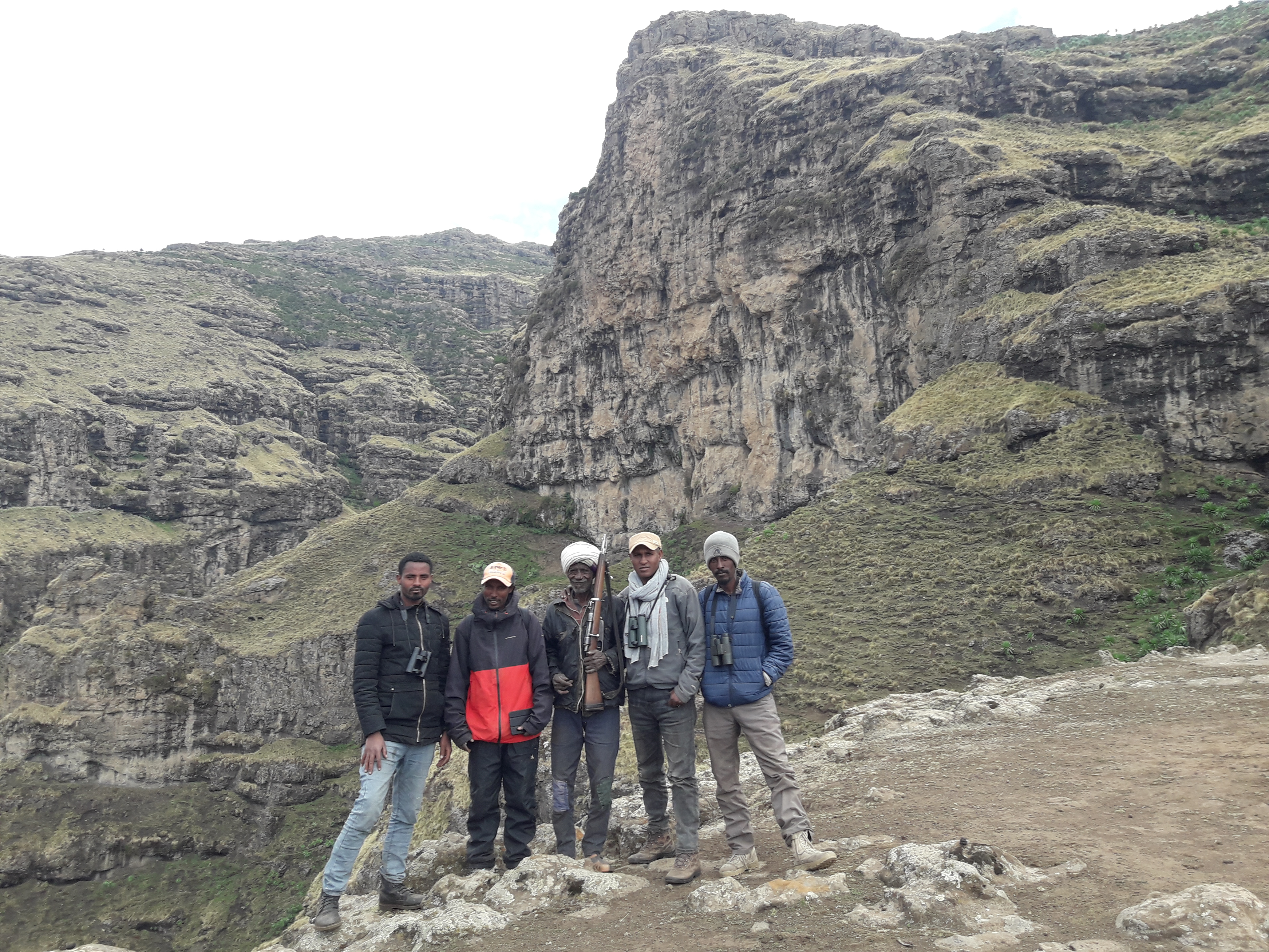 Five men stand on a ridge in front of a mountain landscape